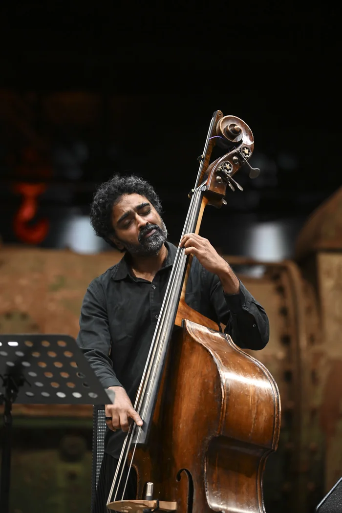 Double bassist Harish Ragshavan plays his instrument with focus. He is wearing a black shirt, his eyes are closed, and he appears deeply immersed in the music. Blurred parts of the industrial hall are visible in the background.