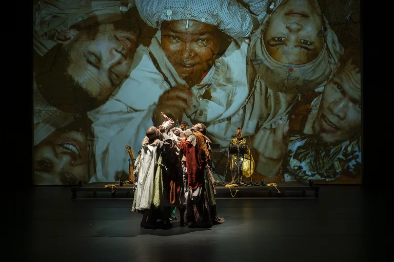 A group of performers in colorful robes stands closely together on stage, with a large projection of faces in the background.