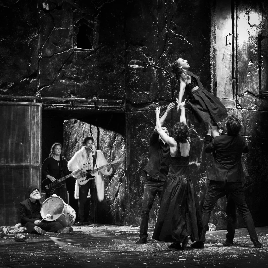 A woman is lifted into the air by several performers on a dark stage. In the background you see a band playing instruments. The photo is in black and white.