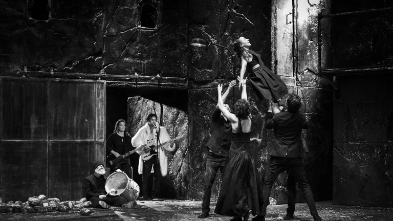 A woman is lifted into the air by several performers on a dark stage. In the background you see a band playing instruments. The photo is in black and white.