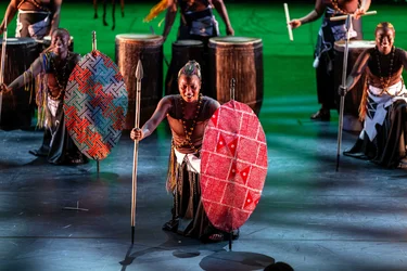 Eine Performerin kniet mit Speer und bemaltem Schild auf der Bühne. Weitere Performerinnen mit Trommeln und Schilden stehen im Hintergrund