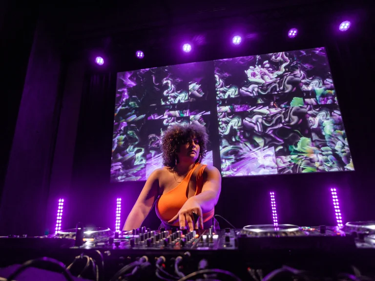 DJ Gîn Bali steht an einem Mischpult auf einer Bühne, beleuchtet von violettem Licht. Im Hintergrund ist ein großer Bildschirm mit abstrakten, psychedelischen Mustern in Violett- und Grüntönen zu sehen.