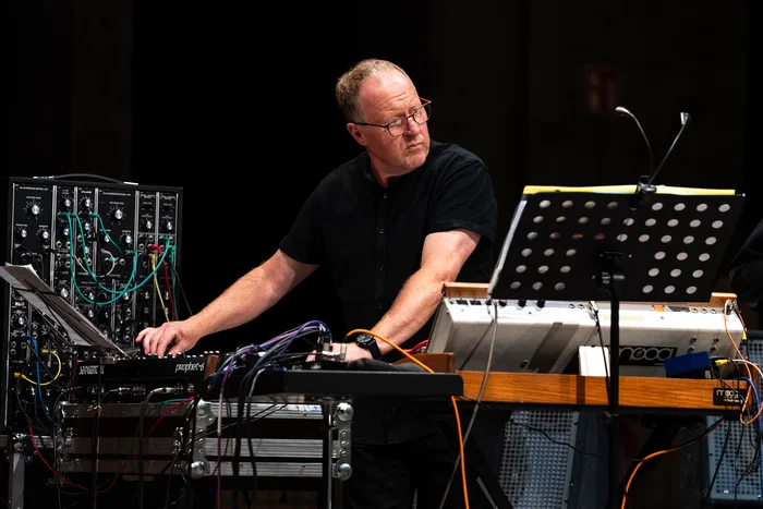 A musician from the Will Gregory Moog Ensemble stands on stage, operating an analog synthesizer while reading sheet music from a stand in front of him. Behind him, a modular synthesizer with numerous colorful cables is visible.