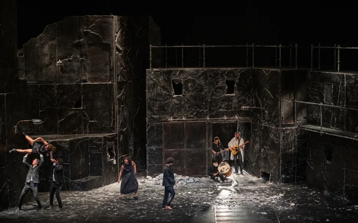 Against a dark, fractured stage backdrop, two performers lift a woman, while others move across the stage. On the right, musicians play guitar and drum.