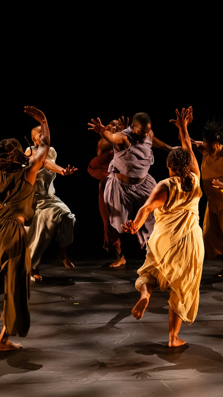 Group of dancers in flowing sleeveless costumes in red, yellow and earthy tones performing a dynamic, synchronized choreography on a dark stage.