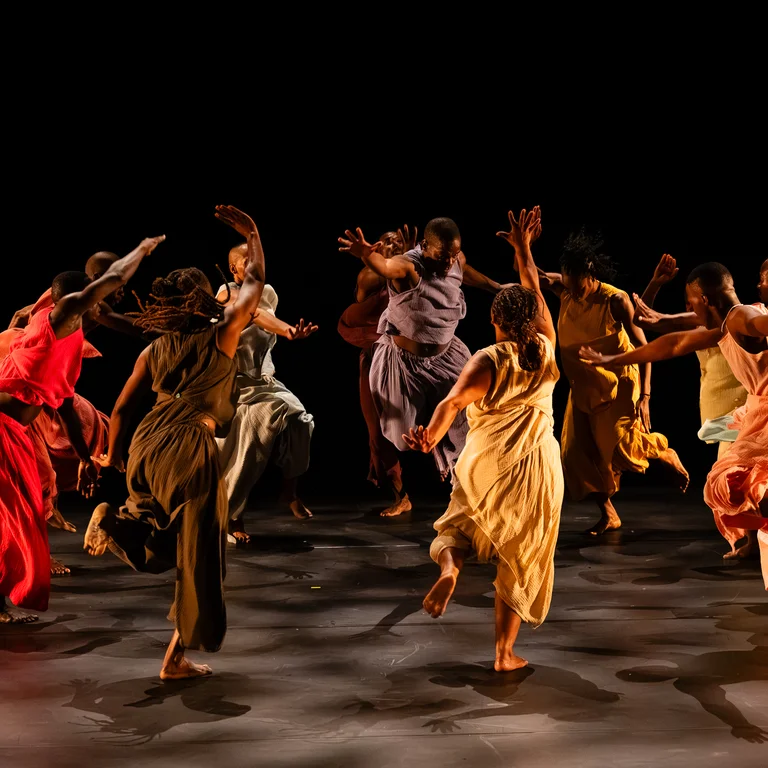 Group of dancers in flowing sleeveless costumes in red, yellow and earthy tones performing a dynamic, synchronized choreography on a dark stage.