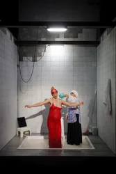 Scene from Balkan Erotic Epic: In a tiled washroom, a woman in a red dress stands in a shallow basin while another woman pours a milk like liquid over her. The scene evokes a ritual cleansing.