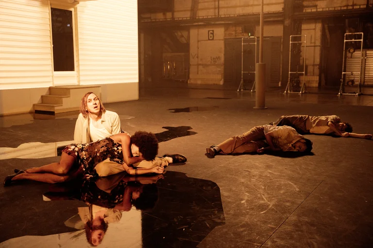 Lars Eidinger is seated on a theater stage in front of a light-colored house façade. Larissa Sirah Herden rests her head on his lap. Her floral dress and afro hairstyle are reflected on a glossy black floor surface. In the background, two other performers lie motionless on the ground. The scene is bathed in warm, orange-colored light.
