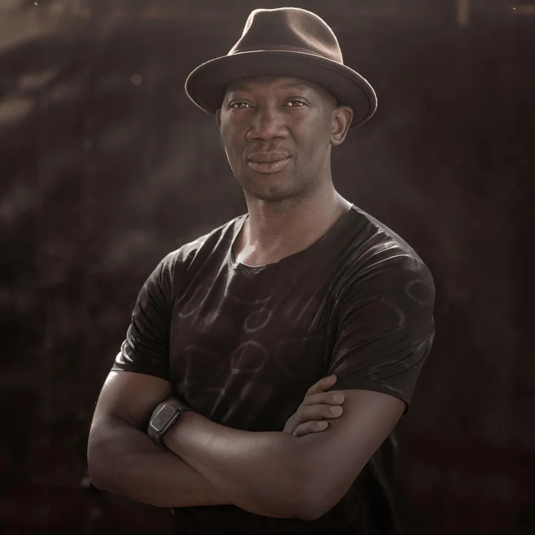 Serge Aimé Coulibaly stands with his arms crossed against a dark background, looking into the camera. He is bathed in sunlight. He is wearing a black T-shirt and a dark hat.