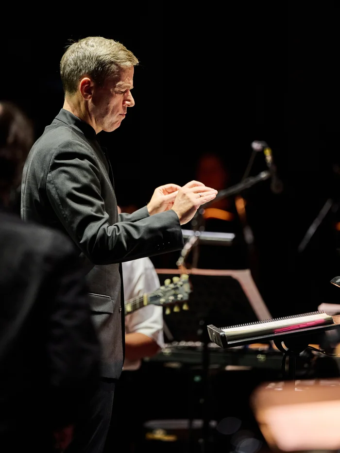 Conductor Florian Helgath conducting on stage, with musicians and instruments blurred in the background.