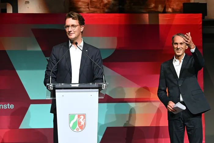 The image shows the opening of the Ruhrtriennale 2024. Minister President Hendrik Wüst is standing at the podium delivering a speech. Next to him, Ivo Van Hove, the artistic director of the Ruhrtriennale, waves to the audience. The background features a colorful stage with the logo of the state of North Rhine-Westphalia. The atmosphere appears festive and formal.