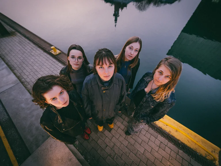 The five members of CATFISH standing next to a shimmering stretch of water. They are all looking slightly upwards at the camera.