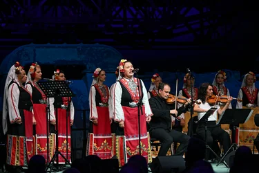 Eine Frau in farbenfroher traditioneller Tracht singt auf der Bühne in ein Mikrofon, umgeben von weiteren Sängerinnen des Frauenchors. Neben ihr spielen zwei Violinist:innen vor einer blau beleuchteten Industriekulisse.