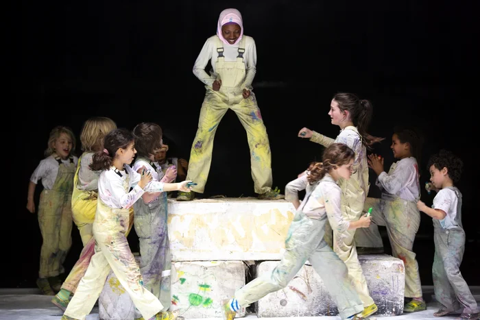 A group of children in painted overalls surrounds another child, who stands confidently on a podium. The children display playful and challenging expressions.