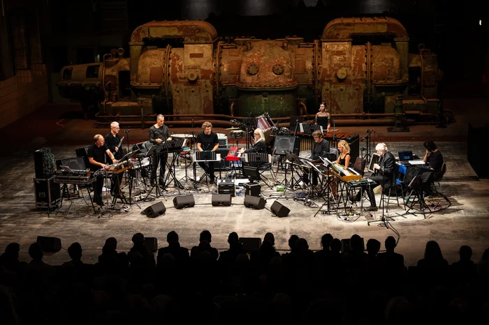 The Will Gregory Moog Ensemble performs in an old industrial hall, with two large, rusty turbines visible in the background. Eleven musicians are seated on stage, playing electronic instruments such as synthesizers, keyboards, and mixing consoles.