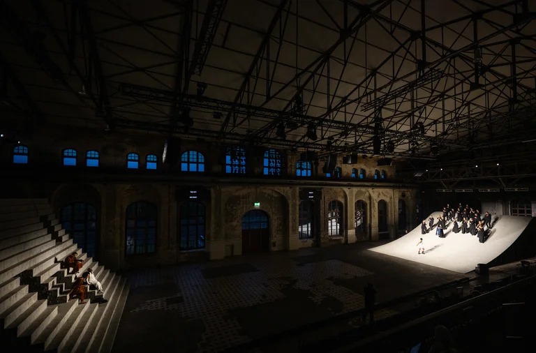 Weite Ansicht einer Theaterbühne in einer großen Industriehalle. Links sitzen drei Darstellende auf einer monumentalen Treppenkonstruktion, rechts steht eine größere Gruppe in schwarzer Kleidung auf einer hell beleuchteten, schrägen Bühne.