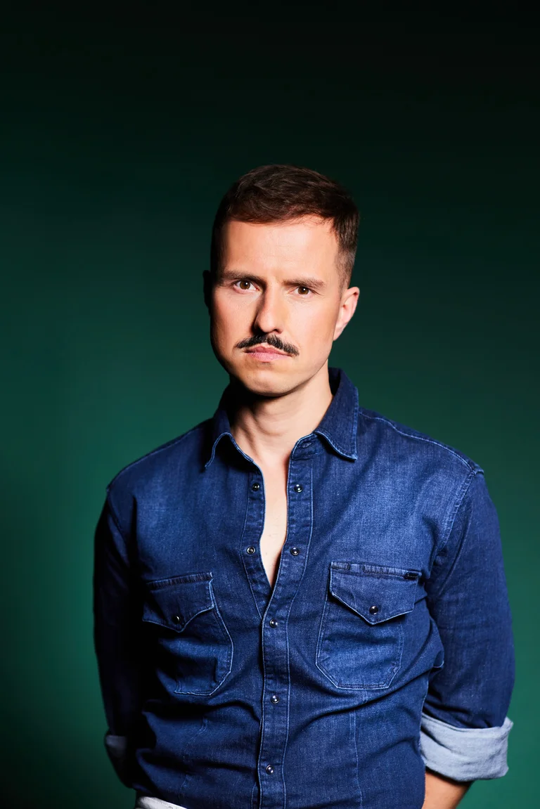 Ein Mann mit Schnurrbart und im Jeanshemd, der Programmdirektor Krystian Lada, posiert vor grünem Hintergrund.