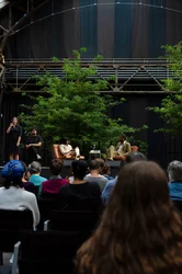 Das Bild zeigt eine Gesprächsrunde im Rahmen der Veranstaltung „Brave New Voices“ in der Jahrhunderthalle Bochum, Teil der Ruhrtriennale. Auf der Bühne sitzen die Moderatorin Fatima Khan und der Schriftsteller Jeremy O. Harris in Sesseln und führen eine Diskussion. Im Vordergrund befindet sich eine Dolmetscherin für Gebärdensprache, die die Konversation übersetzt. Das Publikum sitzt in Reihen vor der Bühne. Die Bühne ist mit zwei großen Bäumen dekoriert, und im Hintergrund ist die industrielle Architektur der Halle mit Stahlträgern sichtbar.
