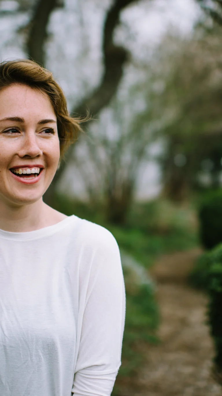 Caroline Shaw standing in front of green hedges and trees. She is wearing a light-colored long-sleeved top and are smiling while looking to the side.