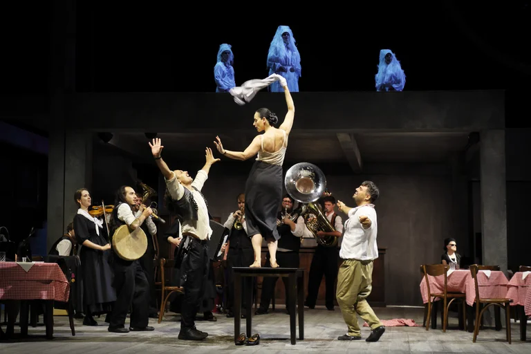 Ensemble scene in a tavern-like set: musicians play, men dance, a woman stands on a table waving a cloth, with white-veiled figures above.