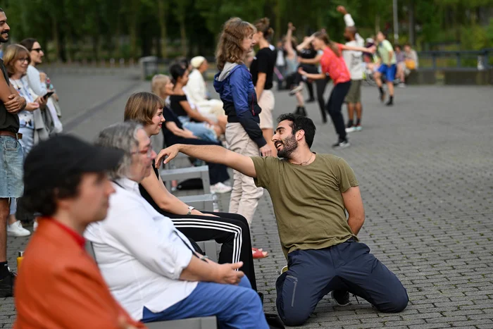 Es ist eine Reihe mit Stühlen mit Publikum zu sehen. Davor tanzen Tänzer:innen zu einer Person auf dem Stuhl. Der Teaser fand am 18.08.2024, auf dem Vorplatz der Jahrhunderhallte in Bochum statt