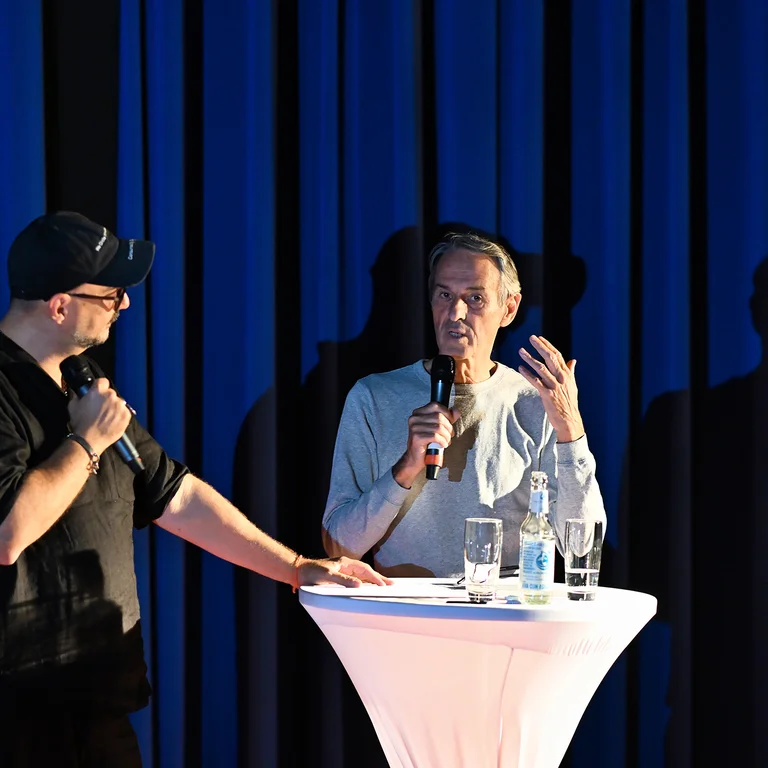 Ivo Van Hove und Regisseur Kirill Serebrennikov stehen an einem weißen Stehtisch vor einem blauen Vorhang in einem Kinosaal.
