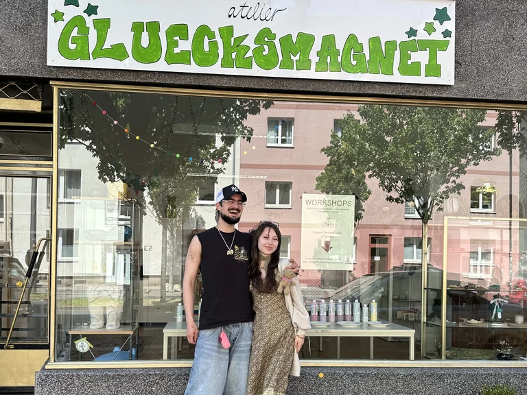 Two people are smiling in front of the shop window of “Atelier Gluecksmagnet” on a city street.