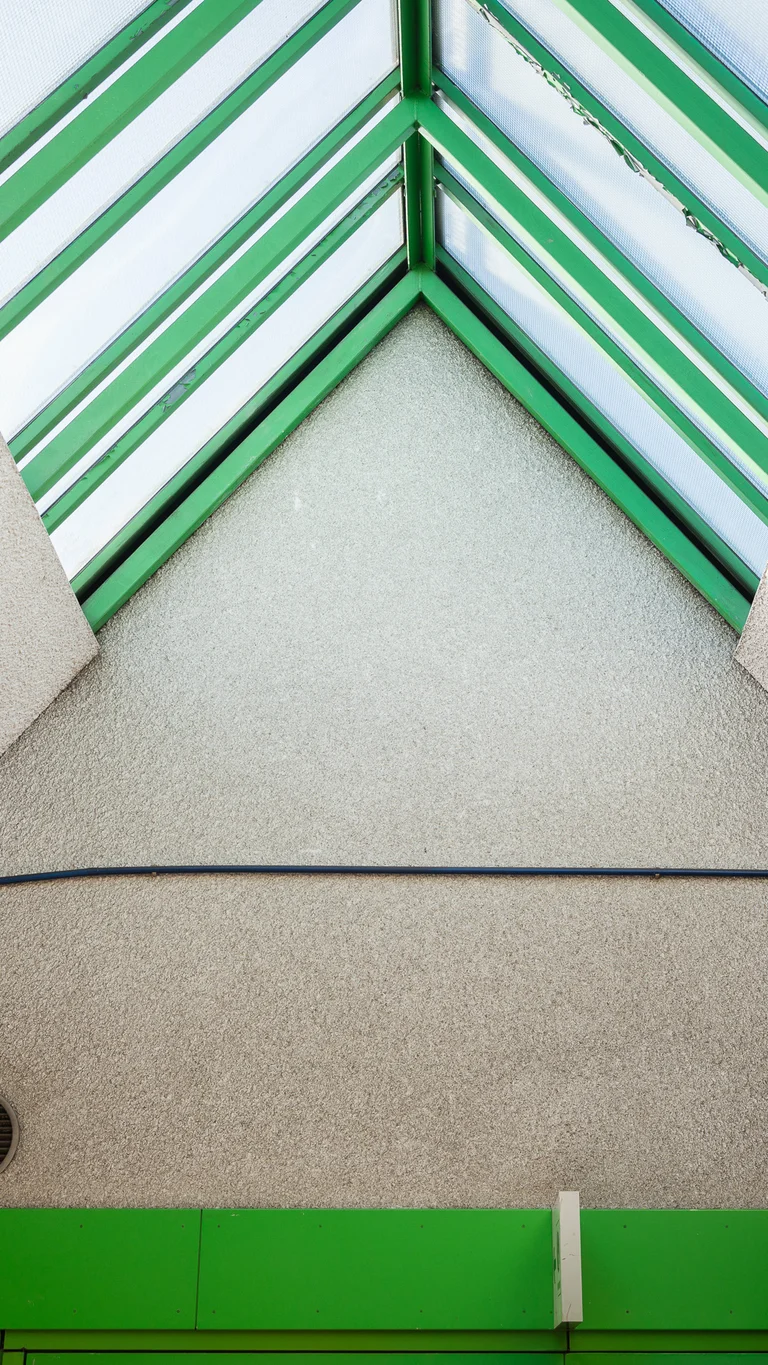 Blick nach oben auf ein spitz zulaufendes, gläsernes Dach aus grünen Streben; an der Wand verlaufen farbige Leitungen und geometrische Wandinstallationen.