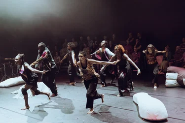 Mehrere Performer:innen tanzen synchron mit nach hinten ausgstreckten Armen über die Bühne. Das Publikum verfolgt die Performance im Hintegrund von der Bühne aus.