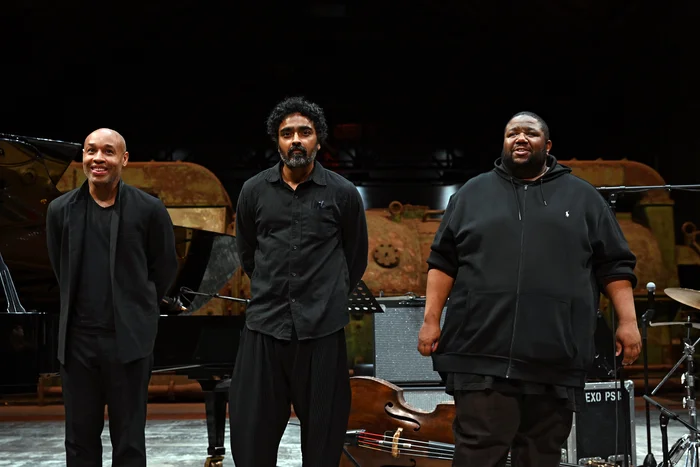 Das Tyshawn Sorey Trio nach dem Auftritt auf der Bühne der Turbinenhalle. Von links nach rechts: Pianist Aaron Diehl, Kontrabassist Harish Ragshavan und Schlagzeuger Tyshawn Sorey stehen nebeneinander und tragen schwarze Kleidung. Im Hintergrund ist die alte, rostige Turbine zu sehen.