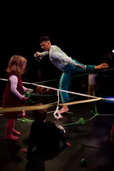 Ein Performer balanciert auf einem Bein, während Kinder ihm zuschauen. Zwischen dem Performer und den zuschauenden Kindern sind Bänder gespannt.