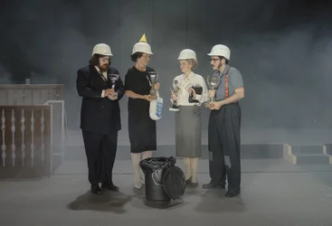 Four performers wearing white hard hats stand on stage holding large silver trophies. A black rubbish bin stands in front of them, and the stage is slightly hazy.