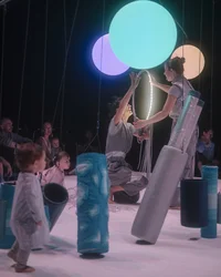 Zwei Performerinnen halten gemeinsam einen leuchtenden Reifen. Um sie herum bewegen sich Kleinkinder zwischen gepolsterten zylindrischen Objekten.