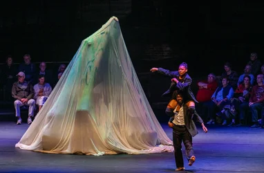 Two performers move acrobatically across the stage, with one seated on the other’s shoulders. Beside them stands a large object draped in light-colored fabric, while the audience sits directly around the performance area.