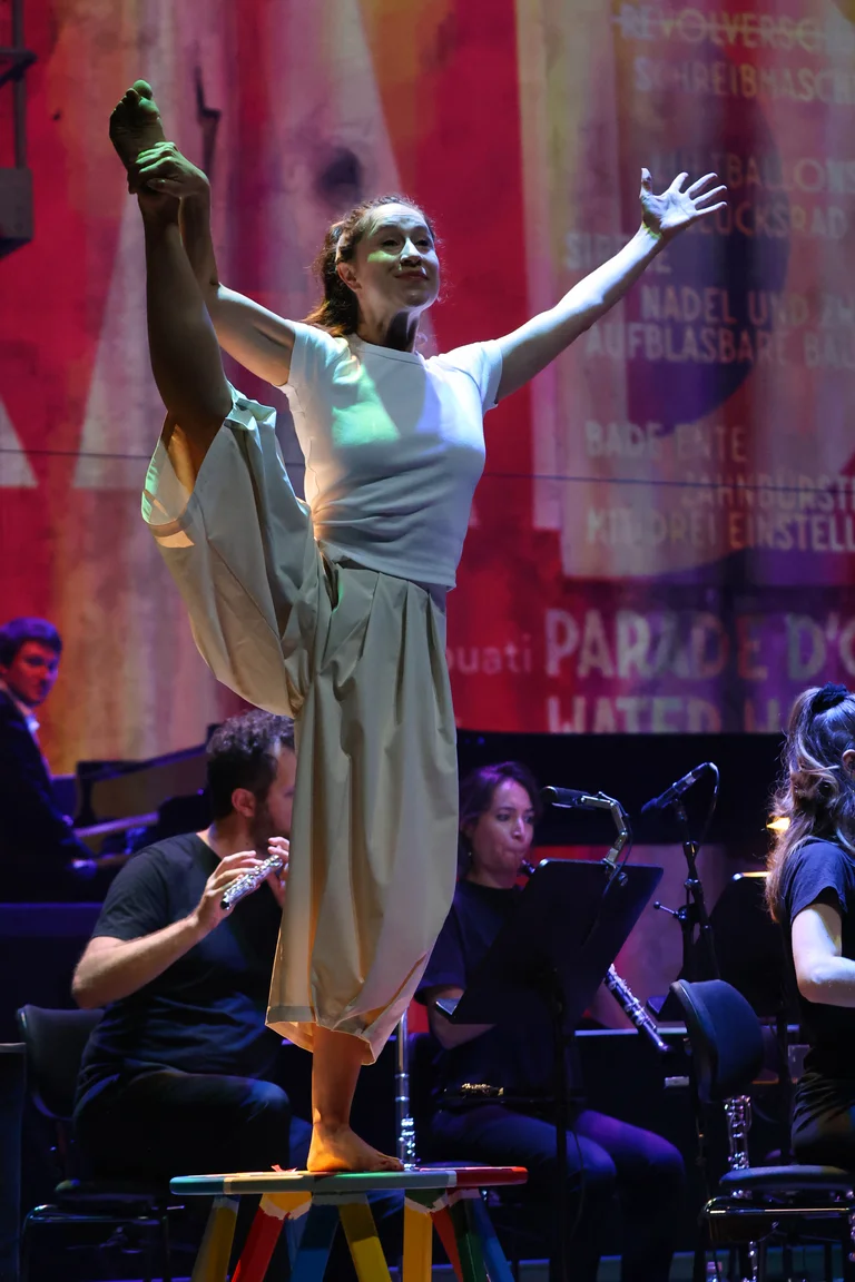 Sarah Silverblatt-Buser balances barefoot on a small colourful bench, raising one leg straight upwards. Her arms are stretched out wide. In the background, musicians from the ensemble Les Apaches ! are seated on stage with their instruments.