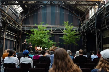 Das Bild zeigt eine Gesprächsrunde im Rahmen der Veranstaltung „Brave New Voices“ in der Jahrhunderthalle Bochum, die Teil der Ruhrtriennale ist. Auf der Bühne, geschmückt mit zwei großen Bäumen, sitzen die Moderatorin Fatima Khan und der Schriftsteller Jeremy O. Harris in Sesseln und führen ein Gespräch. Links steht eine Gebärdensprachdolmetscherin, die die Diskussion übersetzt. Das Publikum sitzt in Reihen und folgt aufmerksam der Diskussion. Der Raum hat eine industrielle Architektur mit Stahlträgern und hohen Decken, was auf die historische Nutzung der Jahrhunderthalle hinweist.
