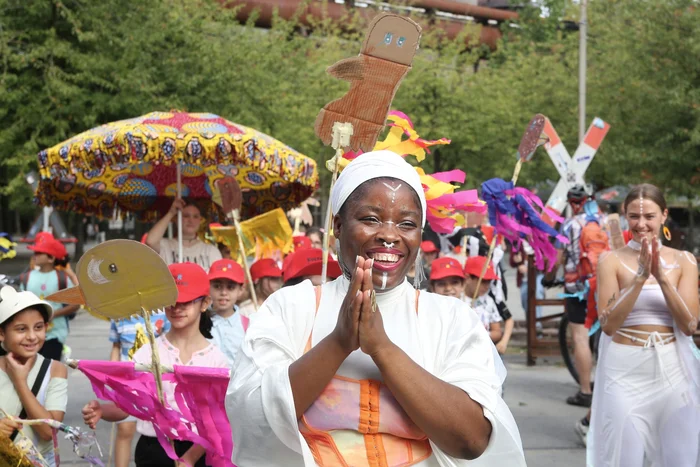 Hiheme performs with a radiant smile is the centerpiece of the 'Celebration Parade'. Children wearing red caps and holding handcrafted figures surround her, with more people and colorful decorations visible in the background.