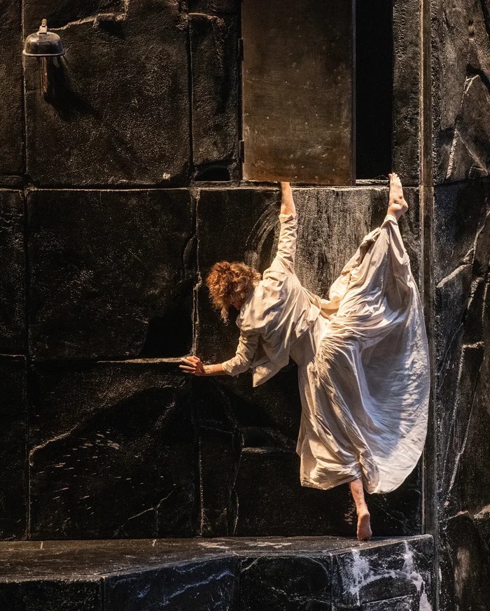 A woman in a long white dress climbs acrobatically on a dark stage wall, holding onto an open hatch.