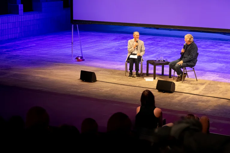 Ivo Van Hove and Will Gregory are sitting on a stage and speaking into microphones.