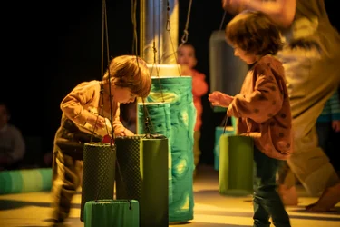 Zwei kleine Kinder stehen in einem abgedunkelten Raum und beschäftigen sich mit hängenden, zylindrischen Objekten in Grün- und Blautönen. Die Objekte sind an Schnüren befestigt und reichen bis auf den Boden. Weitere Kinder und Erwachsene sind unscharf im Hintergrund zu sehen.