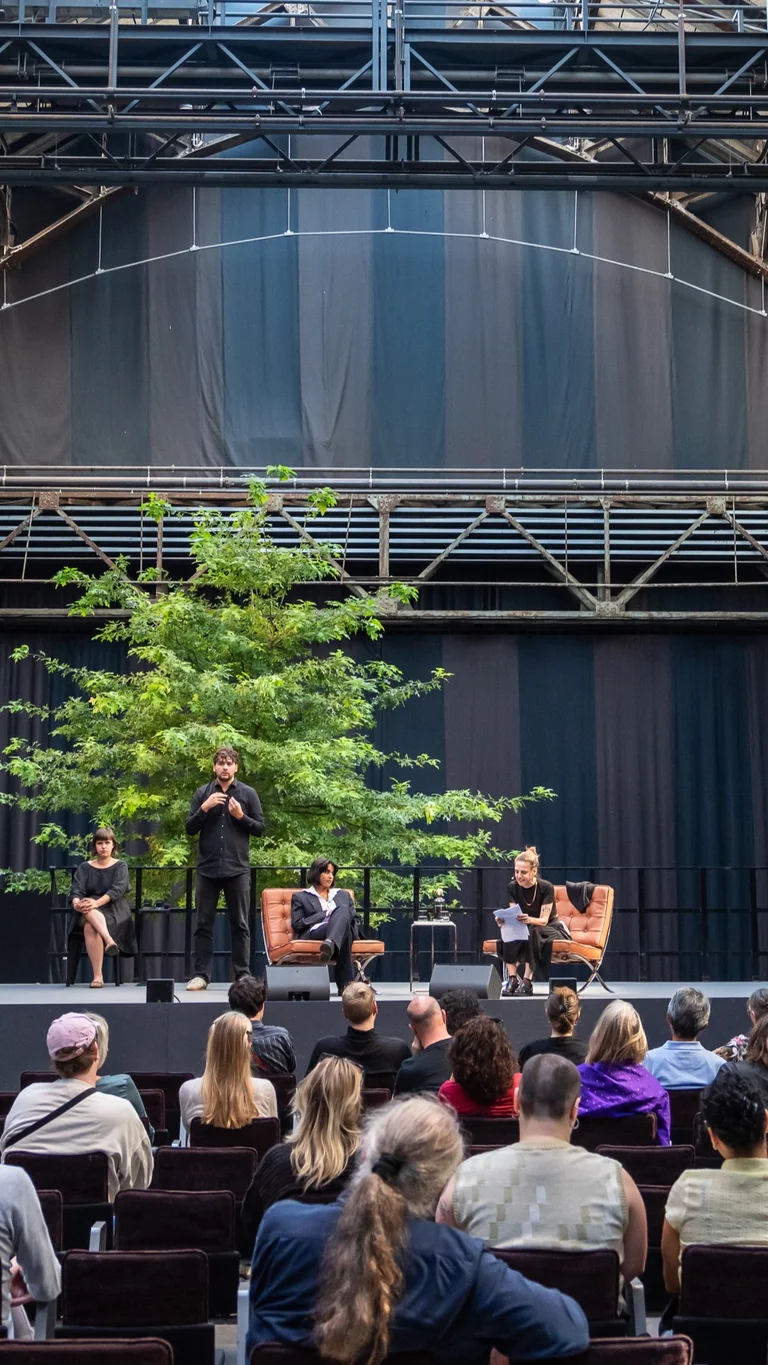 Jahrhunderthalle Bochum: Blick auf die Bühne der Veranstaltung "Brave New Voices". Es sind die Moderation Fatima Khan und Sivan Ben Yishai zu sehen. Daneben sieht man zwei Dolmetscher:innen, die abwechseln in Deutsche Gebärdensprache übersetzen. Im Vordergrund sieht man das Publikum.