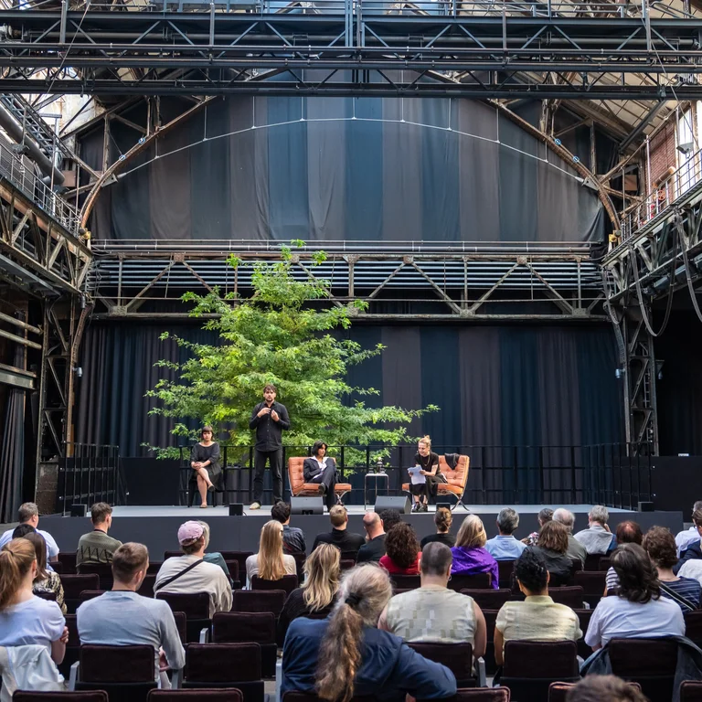 Jahrhunderthalle Bochum: Blick auf die Bühne der Veranstaltung "Brave New Voices". Es sind die Moderation Fatima Khan und Sivan Ben Yishai zu sehen. Daneben sieht man zwei Dolmetscher:innen, die abwechseln in Deutsche Gebärdensprache übersetzen. Im Vordergrund sieht man das Publikum.