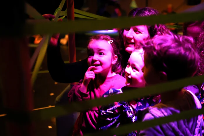 A child and a woman look fascinated at a glowing object behind green tape, while the scene is illuminated in pink-purple light.