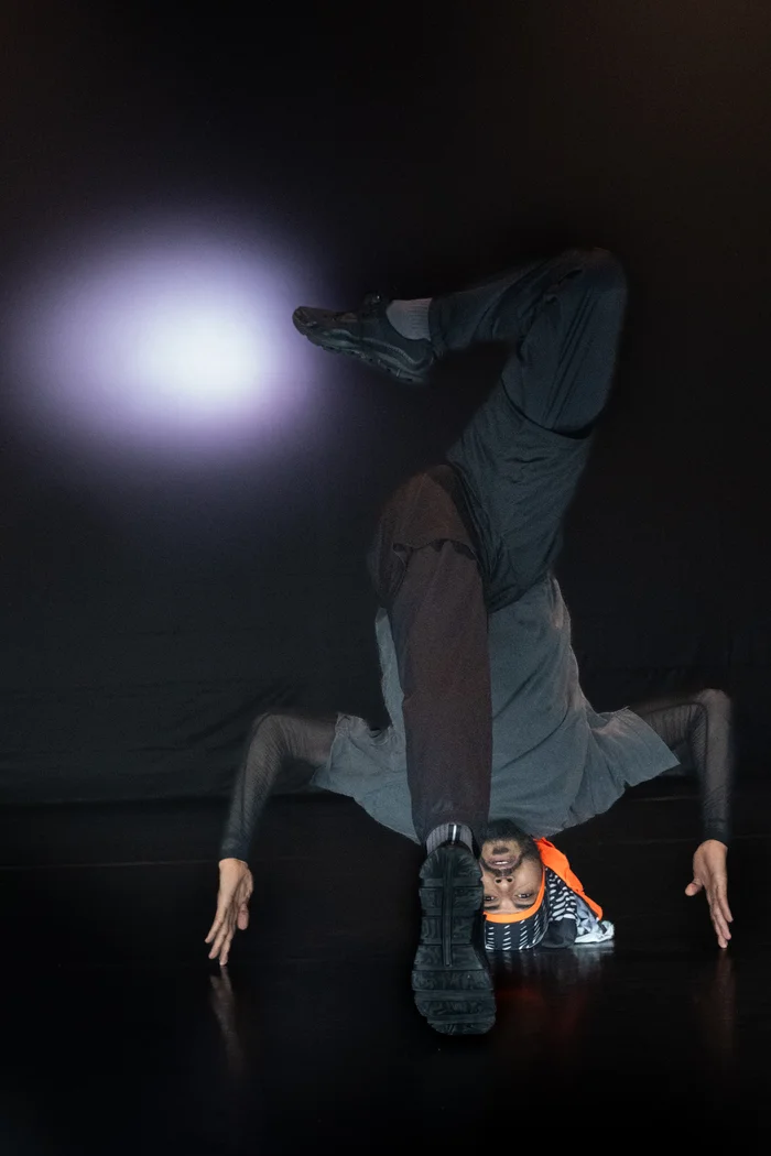 Mohamed Toukabri in an inverted position, supported on arms with head near the floor. An orange bow stands out from the dark outfit.