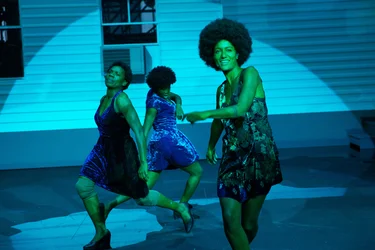 Larissa Sirah Herden and two dancers under colorful stage lighting perform a synchronized dance with raised arms and expressive facial gestures in front of a white house structure.