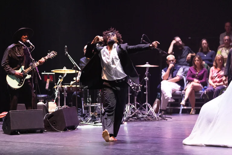 A dancer walks barefoot on stage. He is wearing a black suit, a white shirt, and sunglasses. In the background on the left, a band is playing music. The audience is seated on the right side in the background.