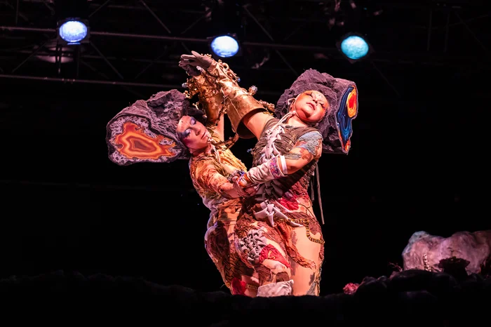 Zwei Performer:innen tanzen eng umschlungen. Sie tragen kunstvoll verzierten Kostüme mit leuchtenden Farben. Beide tragen große, abstrakte Kopfbedeckungen, die wie Gesteinsformationen wirken.