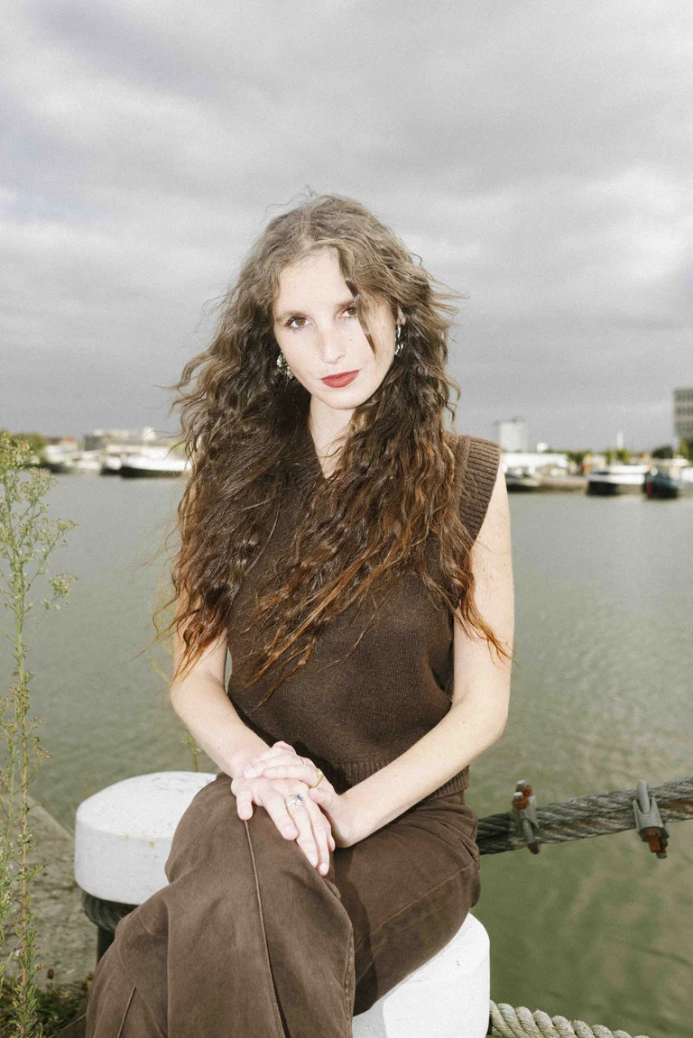 Lisaboa Houbrechts mit langen, lockigen Haaren sitzt an einer Kaimauer vor einem Hafenbecken und blickt direkt in die Kamera. Im Hintergrund sind Wasser, Boote und ein bewölkter Himmel zu sehen.