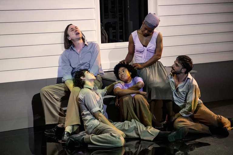 Six performers sit in an emotionally charged scene in front of a light-colored house facade on a theater stage. Lars Eidinger leans against the wall with his shirt open, gazing upward. Next to him, Samuel Planas rests his head on Eidinger's thigh. Sylvie Sanou sits on the steps with her arms crossed, looking thoughtful. Ida Faho gently places her hand on Sanou’s head in a comforting gesture. Marco Labellarte sits on the floor to the right, attentively observing the scene.