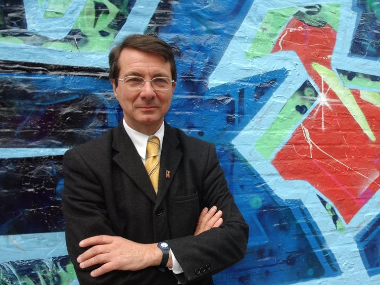 Gerard Mortier in einem Anzug mit Krawatte vor einer Wand mit buntem Graffiti.
